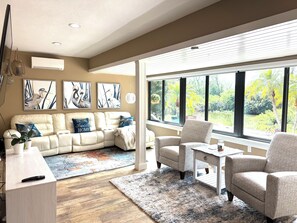 Een woonkamer met een beige bank, een fauteuil met patroon, een salontafel en een groot raam met uitzicht op groen.