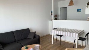 Ein modernes Wohnzimmer mit einem dunkelgrauen Sofa, einem kleinen Holztisch mit Büchern und einer Pflanze, einem gestreiften Tisch mit Stühlen und einer Hängelampe.