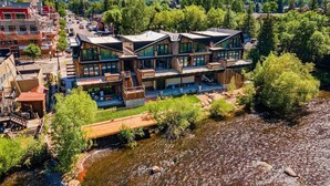 A modern multi-story building with large windows and a wooden facade, located near a river with a mountainous backdrop.