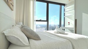 Une chambre moderne avec un grand lit, une vue sur la ville grâce à de grandes fenêtres du sol au plafond, et une table de chevet agrémentée d’un vase de fleurs.