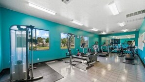 A gym with teal walls, various exercise equipment, and large windows offering views of palm trees and a clear sky.