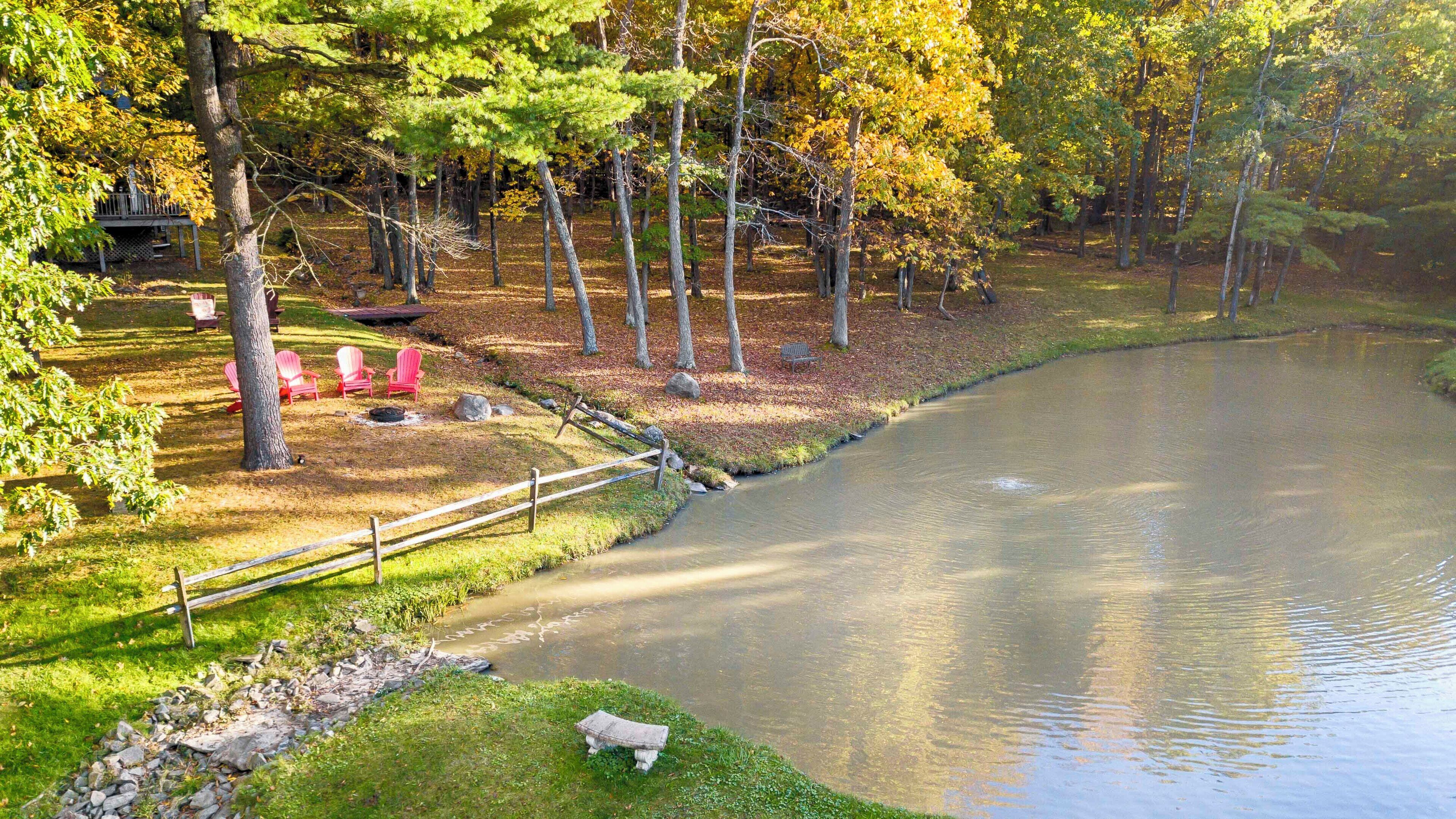 A serene lakeside setting with a wooden deck, red Adirondack chairs, and a stone bench by the water.