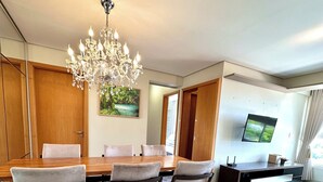 A modern dining area with a wooden table, four upholstered chairs, a chandelier, a painting on the wall, and a television on a stand. - Davisis Luxo Dunas do Jalapão (Palmas)