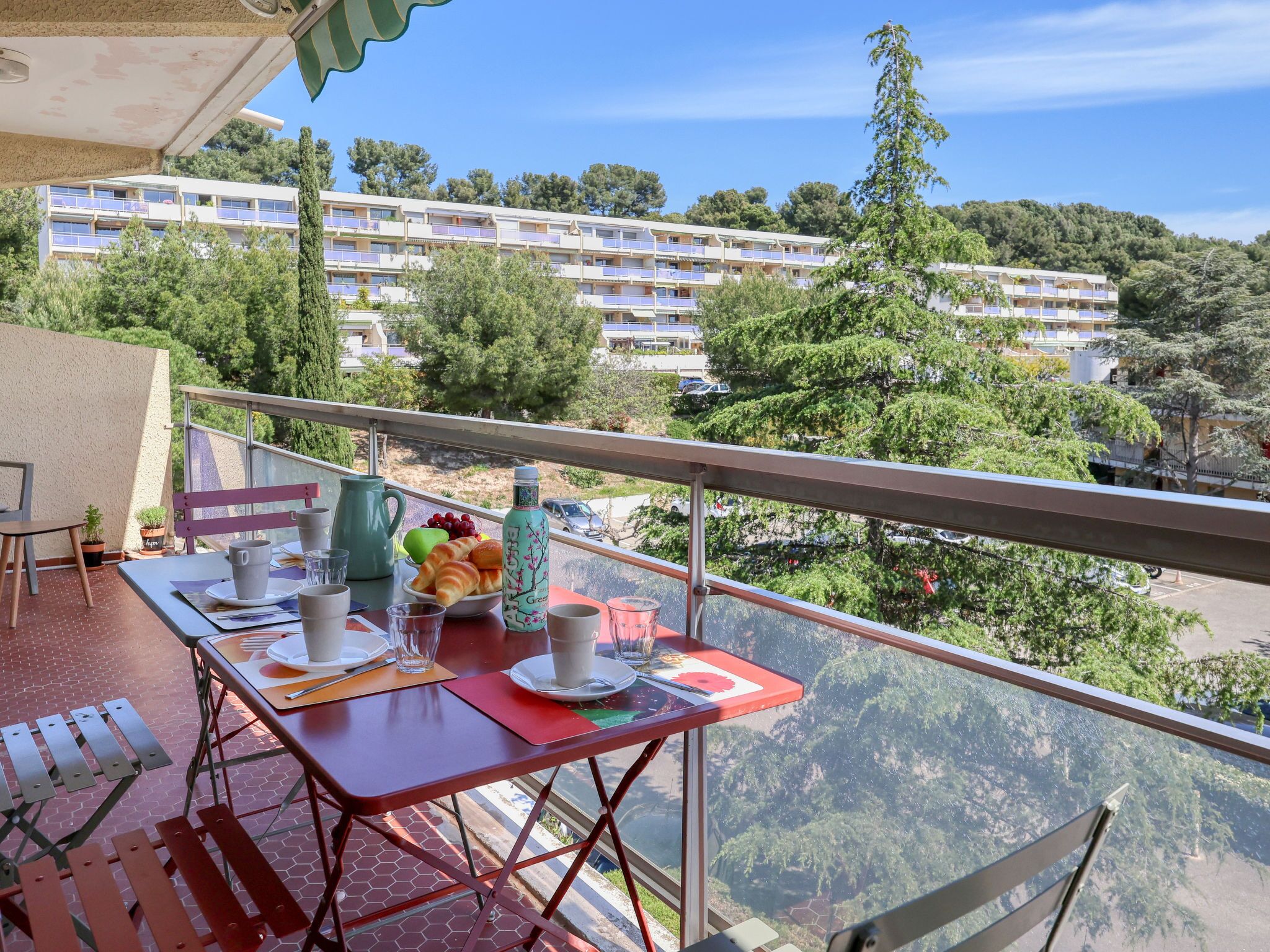 A balcony with a table set for breakfast, including croissants, fruit, and a water bottle. The balcony overlooks a residential area with multi-story buildings and trees.