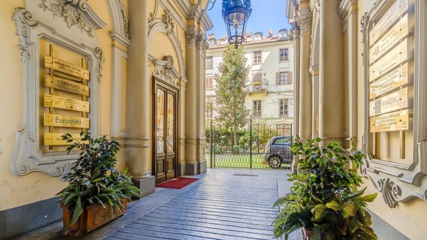 Entrada de hotel con alfombra roja, plantas en macetas y vista a la calle.