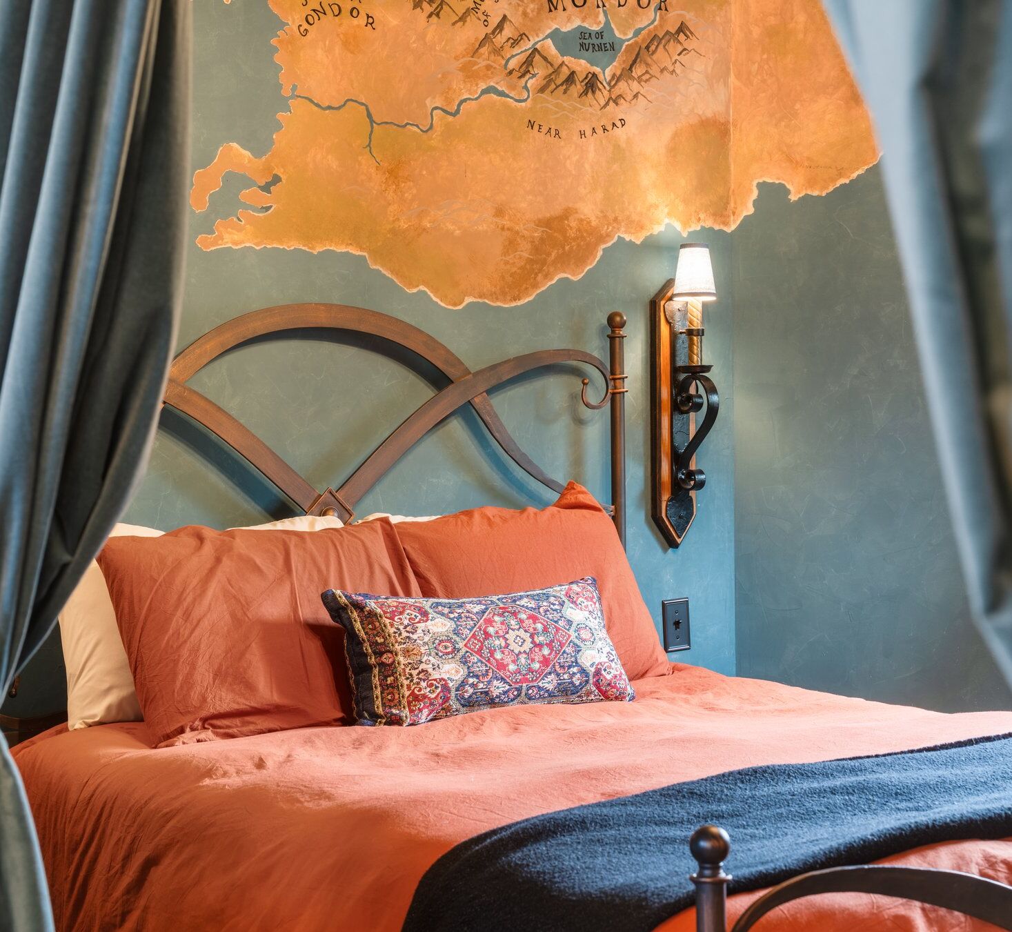 A bed with an orange bedspread and a patterned pillow, a wall-mounted lamp, and a map of Middle-earth on the wall.