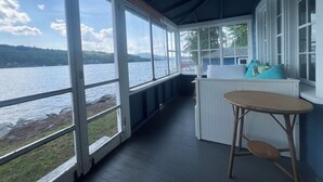 A covered porch with large windows overlooking a lake, a white bench, and a small table.