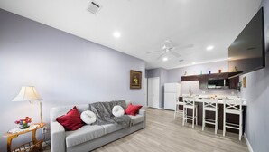 Una sala moderna con un sofá gris, cojines rojos y blancos, una mesita auxiliar con una lámpara y una mesa de centro de madera. Se ve una zona de cocina con taburetes blancos y una televisión de pantalla plana al fondo.