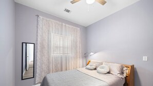 Un dormitorio con cama, ventilador de techo, ventana con cortinas translúcidas y espejo en la pared.