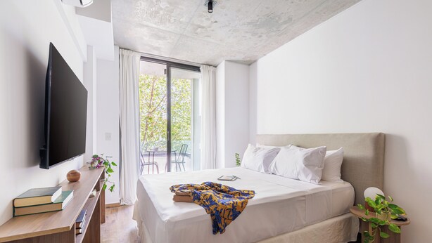 A modern bedroom with a large bed, a flat-screen TV, a bedside table with books, and a view of the outdoors. - Hestia at Thames 2200 (Buenos Aires)