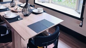 A dining area with a wooden table set for two, a large window with a view of trees, and hanging pendant lights. - Dual Peaks Gem $1M Sharp Views /HOT TUB & WiFi (Tuckasegee)
