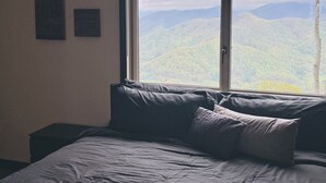 A bedroom with a large window offering a mountain view. There is a bed with grey bedding and a decorative pillow. Two framed items are visible on the wall. - Dual Peaks Gem $1M Sharp Views /HOT TUB & WiFi (Tuckasegee)