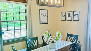 Un coin repas avec une table en bois, des chaises noires, un chemin de table blanc et un vase avec des fleurs. Des tableaux encadrés ornent le mur, et une fenêtre est équipée de stores.