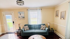 Un salon avec un canapé vert, une table basse et une table à manger avec des chaises. On y trouve une porte donnant sur l’extérieur, un luminaire au plafond et deux objets décoratifs fixés au mur.