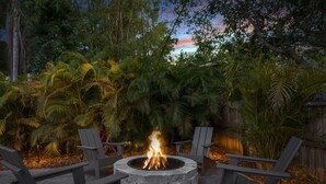 Un pozo de fuego circular rodeado de cuatro sillas Adirondack, en un jardín con abundante follaje y una valla de madera al fondo.