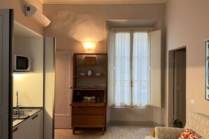 A compact living space with a kitchenette, a built-in shelf unit, a window with sheer curtains, and a patterned sofa. - Charming One-Bedroom Apartment in the Heart of Florence (Firenze)