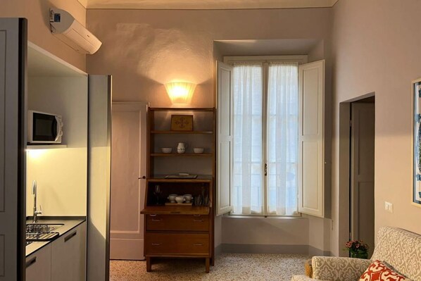 Un espace de vie compact comprenant un coin cuisine, une étagère intégrée, une fenêtre avec des rideaux légers et un canapé à motifs.