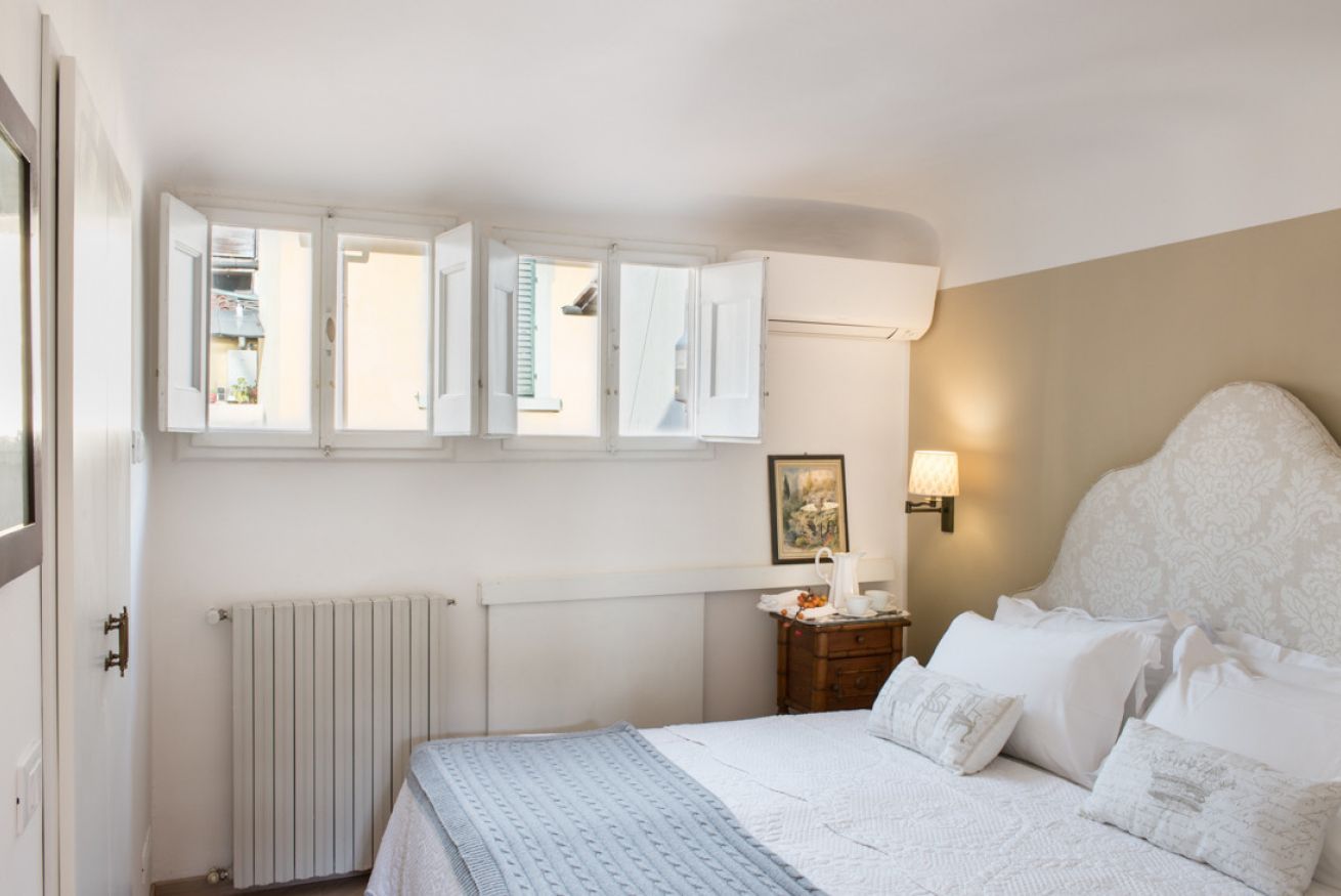 A bedroom with a bed, a nightstand, a radiator, and a window with open shutters.