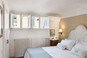Un dormitorio con cama, mesita de noche, radiador y ventana con contraventanas abiertas.