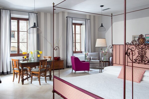 Ein Hotelzimmer mit Himmelbett, Esstisch mit Stühlen, einer Couch und einem Sessel. Es gibt Fenster mit Vorhängen, eine Lampe und eine Vase mit Blumen.