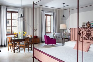 A hotel room with a four-poster bed, a dining table with chairs, a sofa, and a chair. There are windows with curtains, a lamp, and a vase with flowers.