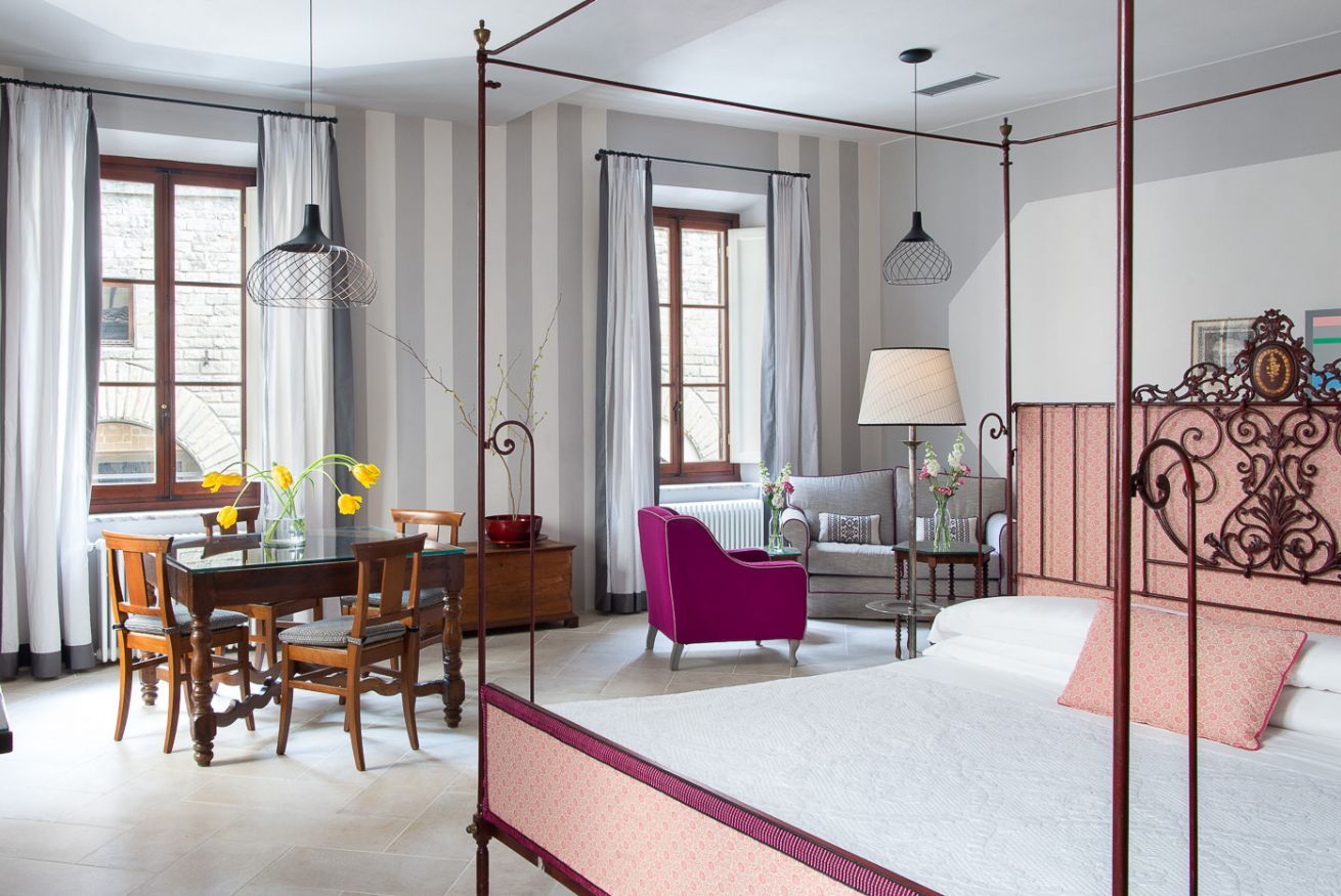 A hotel room with a four-poster bed, a dining table with chairs, a sofa, and a chair. There are windows with curtains, a lamp, and a vase with flowers.