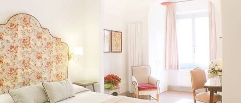 Un dormitorio con cabecera de flores, una cama con almohadas, una silla, una mesa con flores y vistas al exterior a través de una ventana.