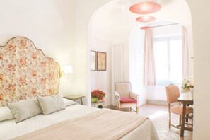 Une chambre à coucher avec une tête de lit à motifs floraux, un lit avec des oreillers, une chaise, une table ornée de fleurs et une vue sur l’extérieur par une fenêtre.