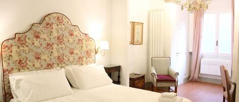 A neatly made bed with white linens and a floral headboard, a chair with a red cushion, and a chandelier.