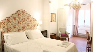 Una cama bien tendida con sábanas blancas y un cabecero floral, una silla con cojín rojo y una araña.