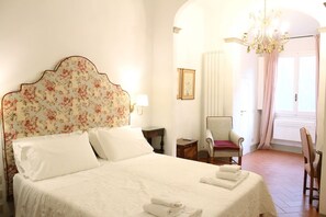 Ein ordentlich bezogenes Bett mit weißen Leinen und einem blumengemusterten Kopfteil, ein Sessel mit rotem Kissen und eine Chandelier.