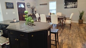 Una isla de cocina con encimera negra y un arreglo decorativo de plantas verdes. Hay luces colgantes sobre la isla, y un área de comedor con sillas y una mesa al fondo.