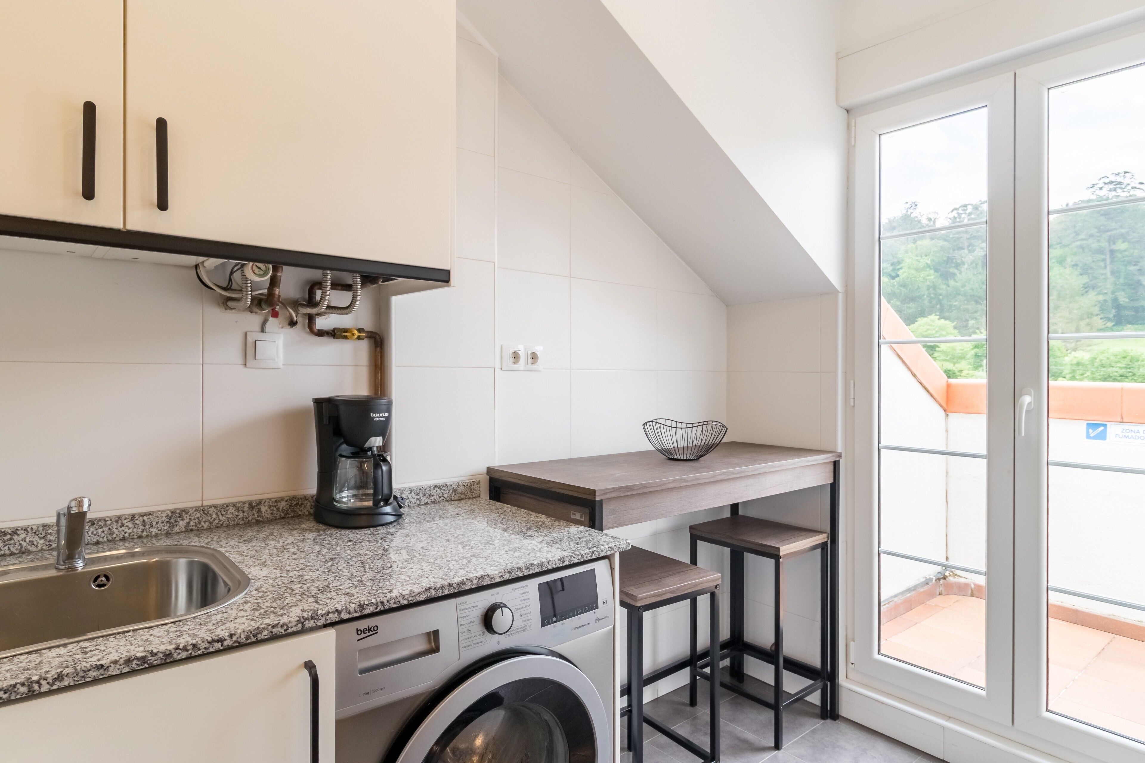 Cocina compacta con lavadora, cafetera y vistas al exterior a través de un ventanal corredero.