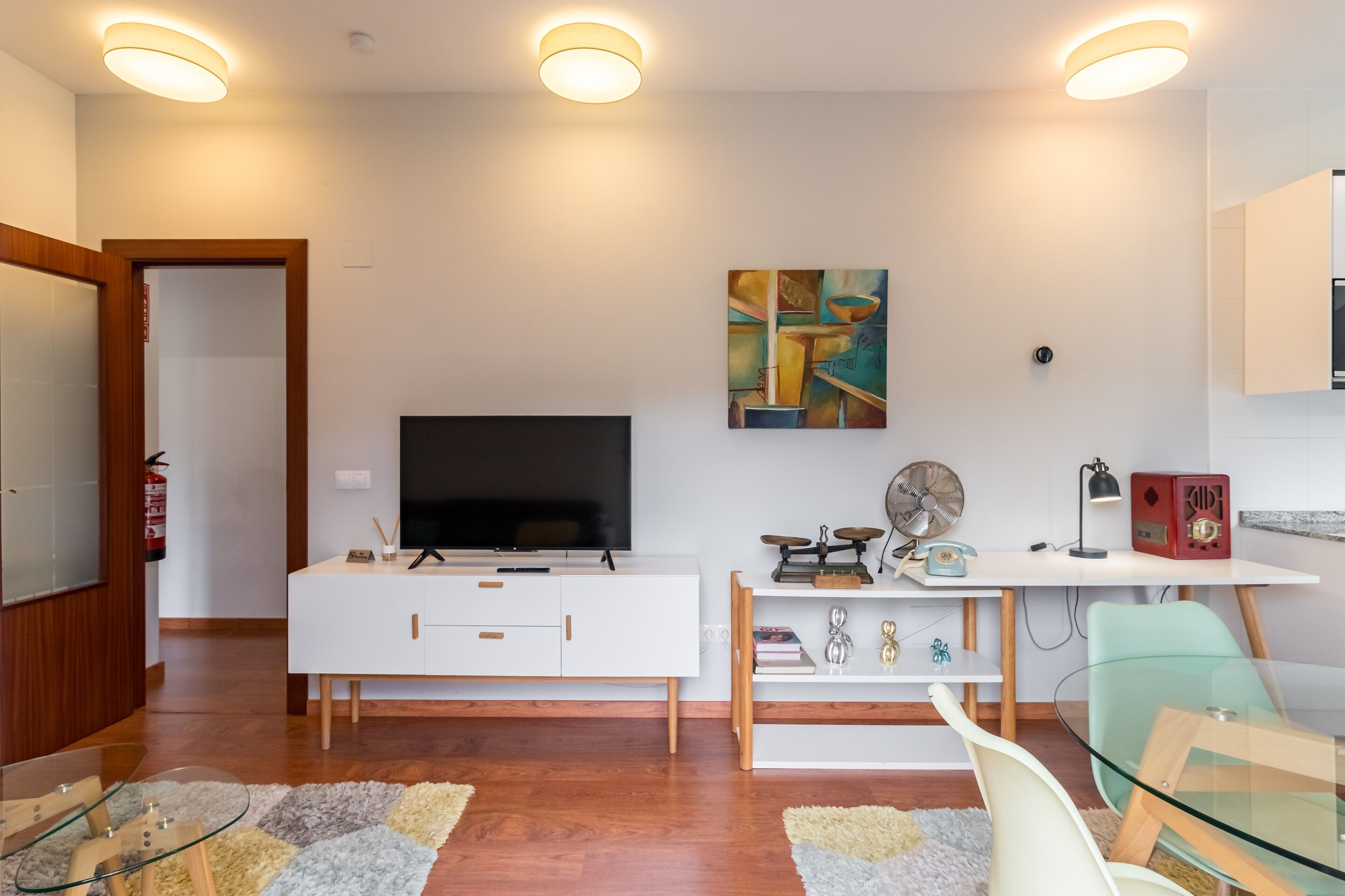 A modern living room with a flat-screen TV, white furniture, a painting, and a desk with a vintage radio.