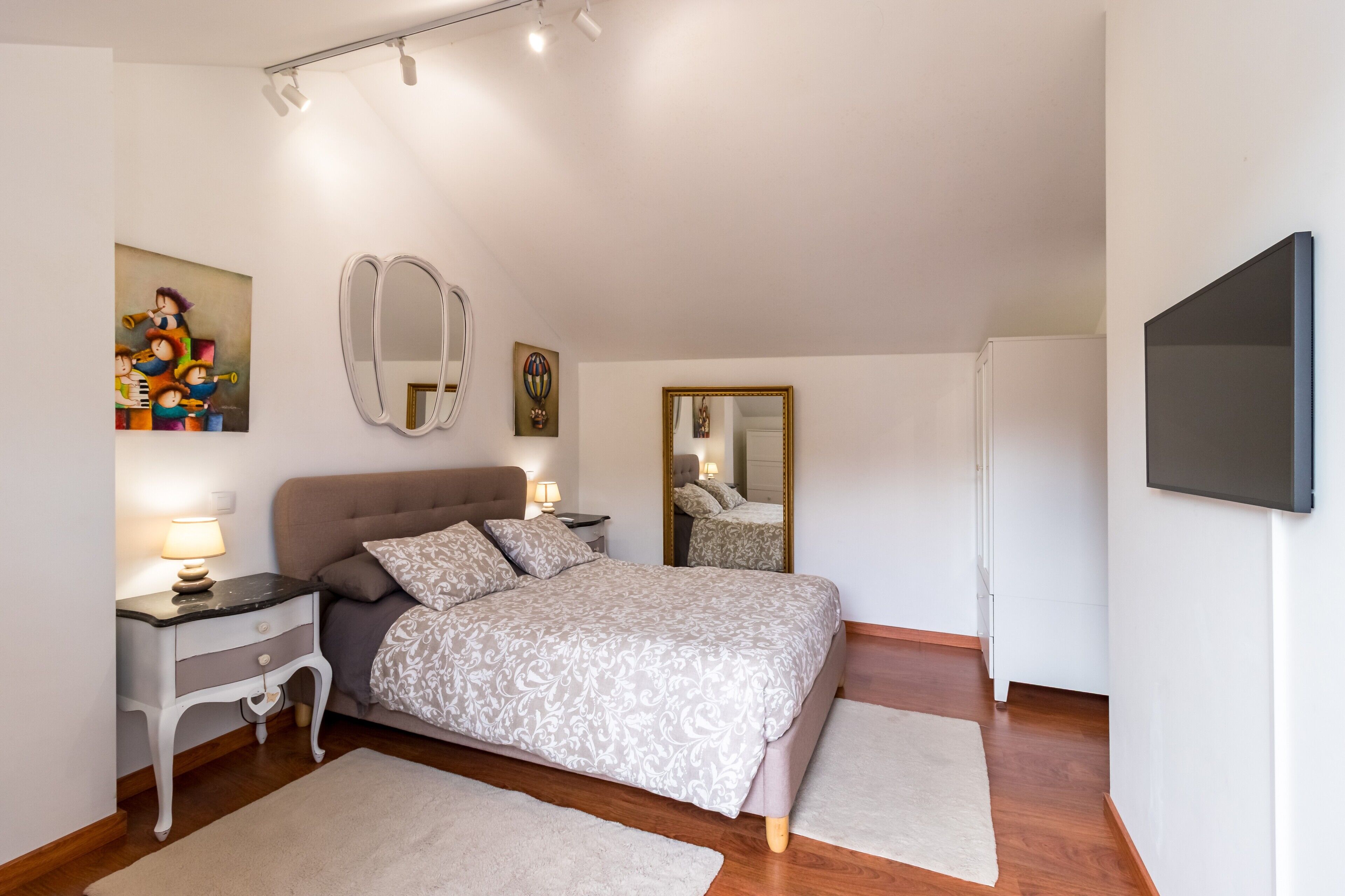 Een slaapkamer met een bed, nachtkastjes, een spiegel, een televisie en kunst aan de wanden.