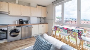 A compact kitchen with modern appliances, a dining area with a colorful tablecloth, and a view of the city through large windows. - Apartment 'Luminoso' with Mountain View, Private Terrace and Wi-Fi (Renedo de Piélagos)