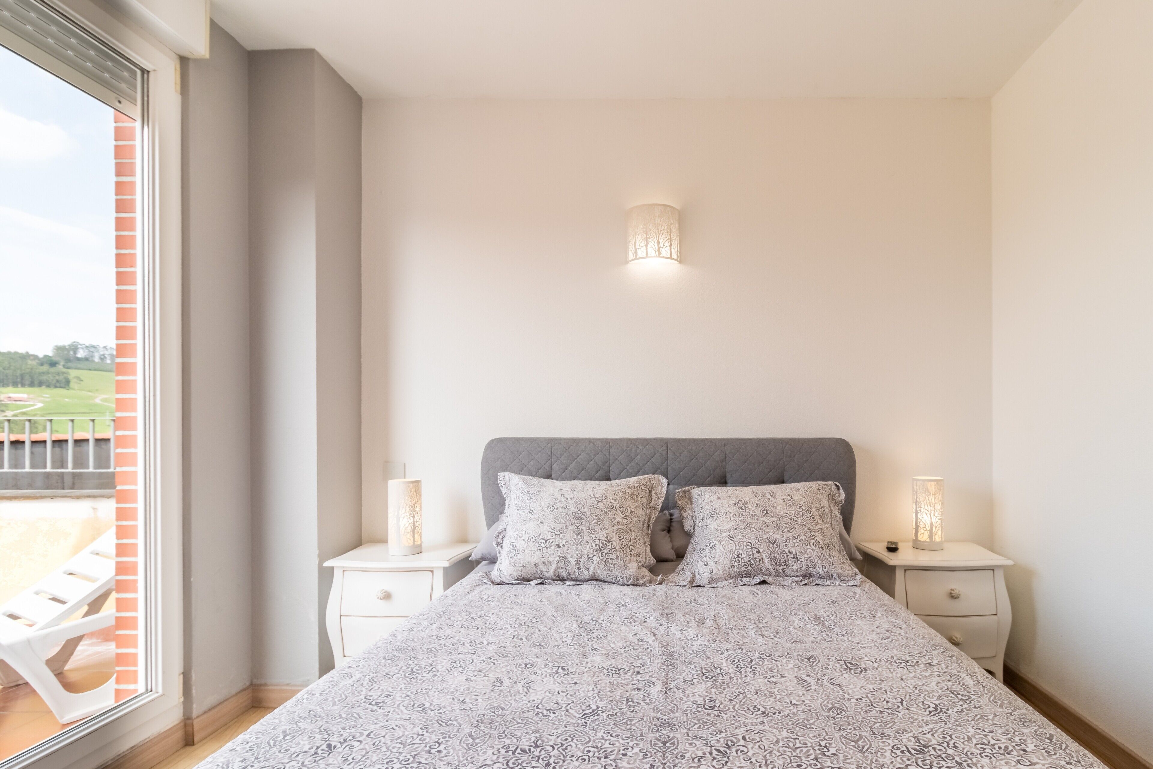 Un dormitorio con una cama, dos mesitas de noche, un pequeño mueble y una ventana con persianas, con vistas a un paisaje verde.
