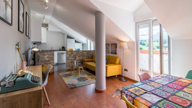 Un dormitorio estilo loft moderno con un ventanal grande, un sofá amarillo, un escritorio con máquina de escribir y una colcha colorida.