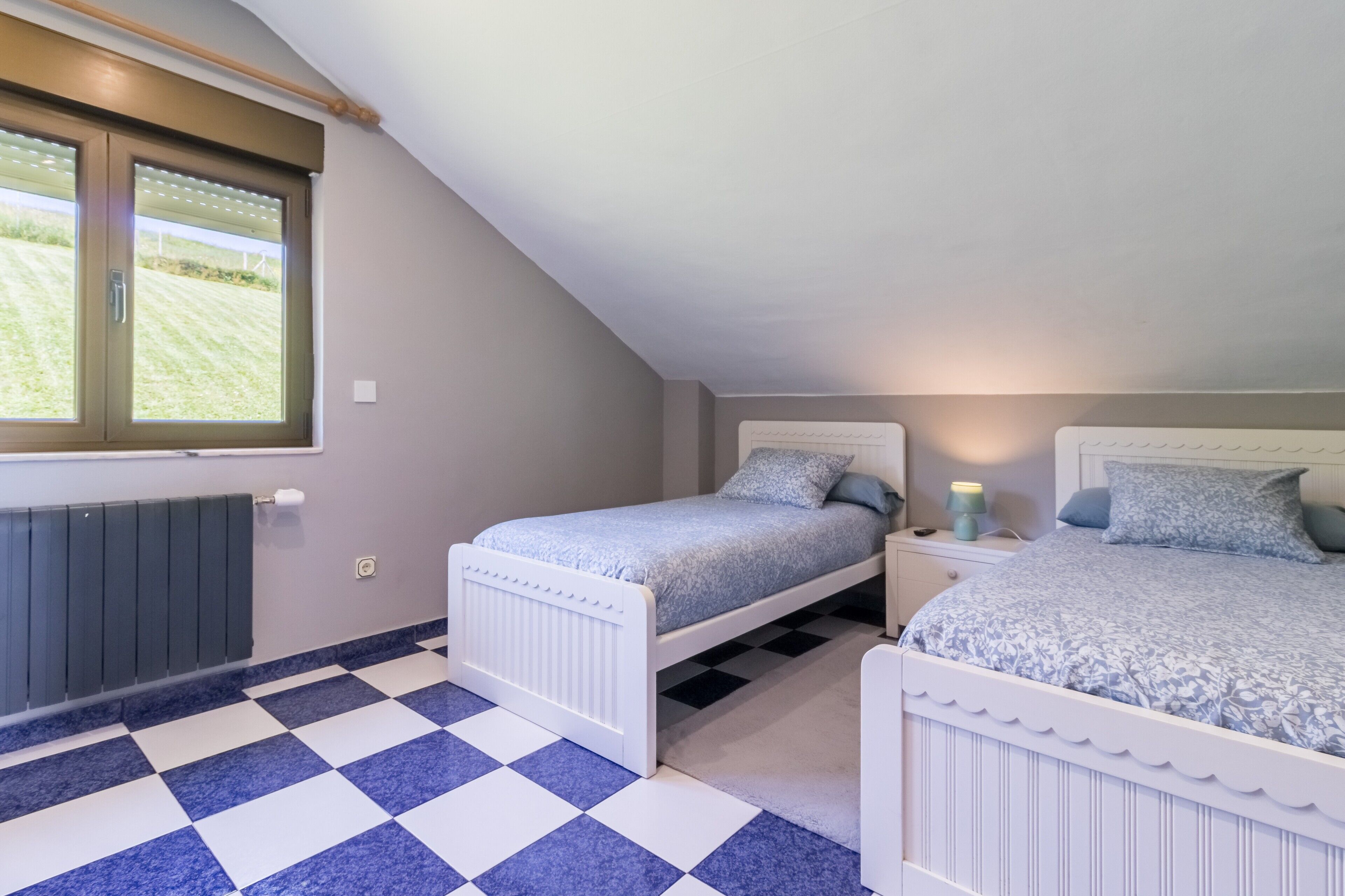 Une chambre avec deux lits, un sol à carreaux, une fenêtre donnant sur un paysage agréable et un radiateur.