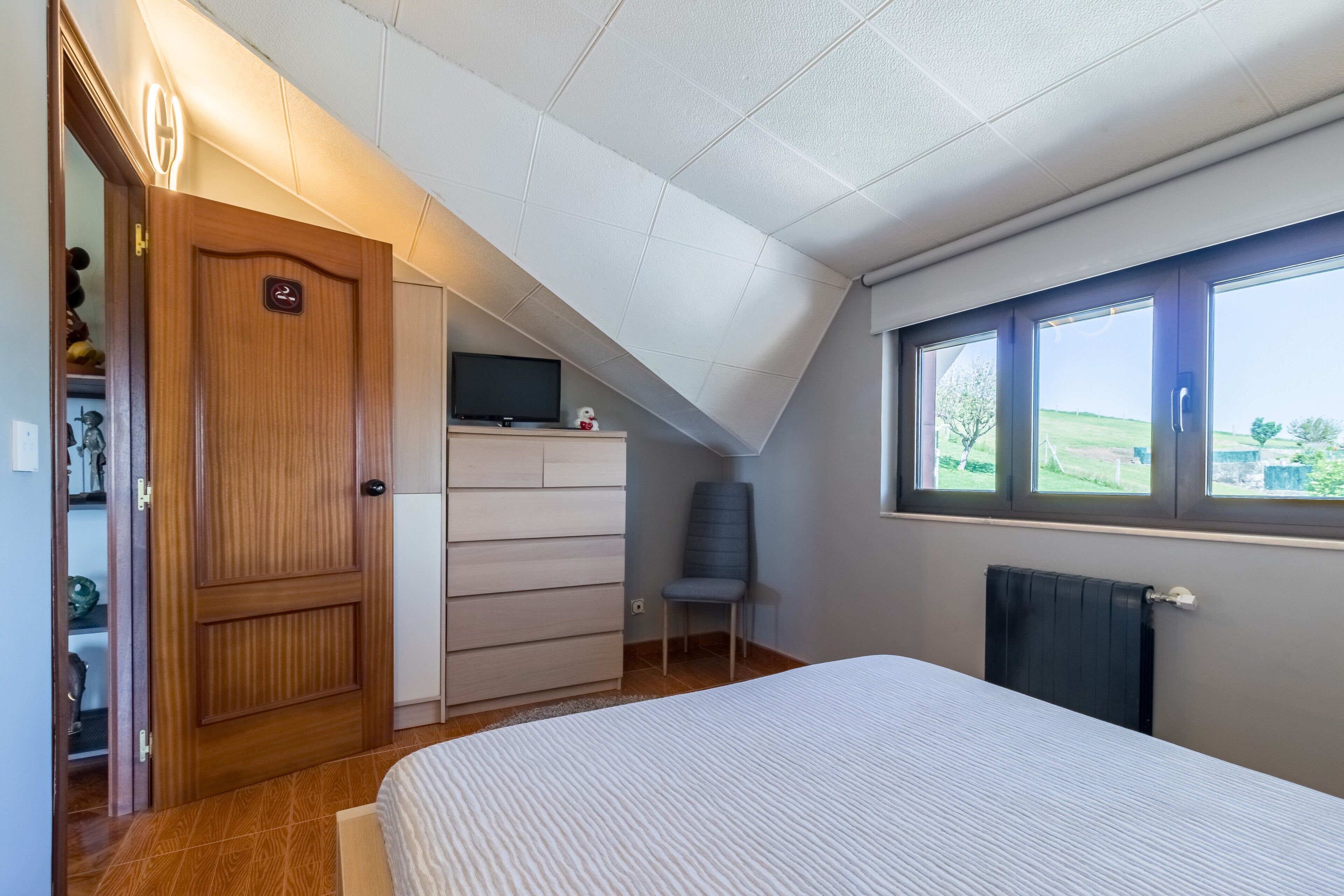 A bedroom with a sloped ceiling, a bed, a dresser, a chair, and a window with a view of greenery.