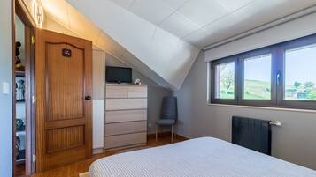 A bedroom with a sloped ceiling, a bed, a dresser, a chair, and a window with a view of greenery.