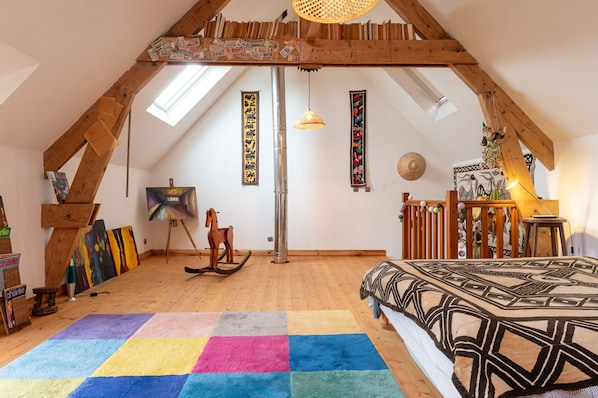 Une chambre à coucher aménagée dans un grenier, avec un plafond en pente, des poutres en bois, un tapis coloré, une couvre-lit à motifs, un cheval à bascule et une grande fenêtre.