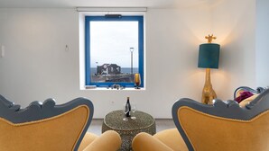 A room with a view of a coastal town, a small round table with a bottle and glasses, two chairs with a unique design, and a lamp with a golden figure.