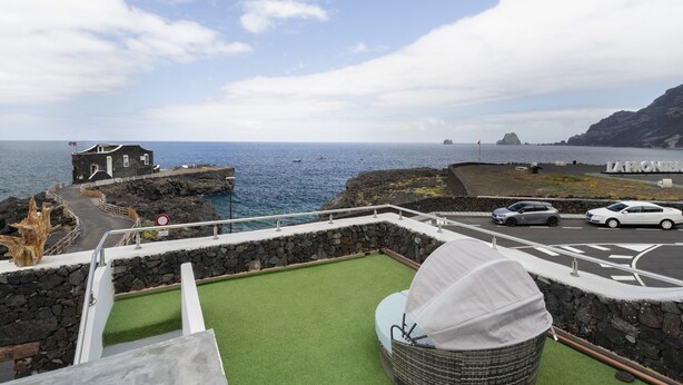A balcony with a view of the sea, a stone wall, a white car, and a building with a flag.