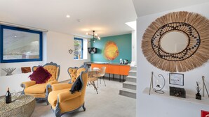 A modern living room with a large painting, a round mirror, and a dining area with orange cabinetry.