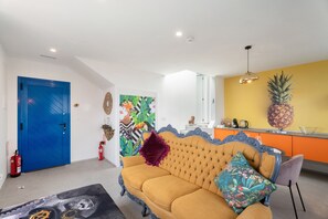 Una sala de estar con un sofá, una mesa de centro, una puerta azul y una zona de cocina con una decoración de piña.