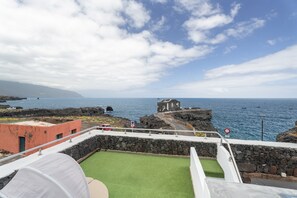 Vista desde un balcón sobre una zona costera con un muro de piedra, un edificio y el mar.