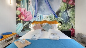 Ein Schlafzimmer mit Wandgemälde von Blumen und Mond, ein Bett mit weißer Bettwäsche, ein Nachttisch aus Holz und ein Ventilator.