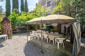 An outdoor dining area with a table and chairs under a canopy, surrounded by trees and a stone building.