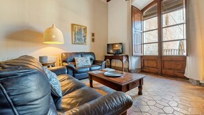 Una sala de estar con sofás de cuero, una mesa de centro de madera, una lámpara colgante, un televisor y un ventanal con vistas a un edificio de ladrillo.
